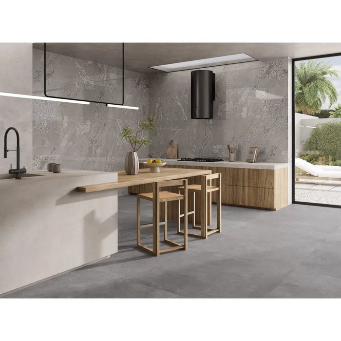 Modern kitchen interior with a light wood island table and three matching bar stools, set against gray stone-look tile walls and floor. A black range hood, pendant lights, and large glass doors showing an outdoor patio are visible.
