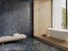 A modern bathroom interior featuring large slate-look tiles in blue, gray, and gold veining on the wall and floor, a light wood platform, a white freestanding bathtub, and a minimalist bench with folded towels.